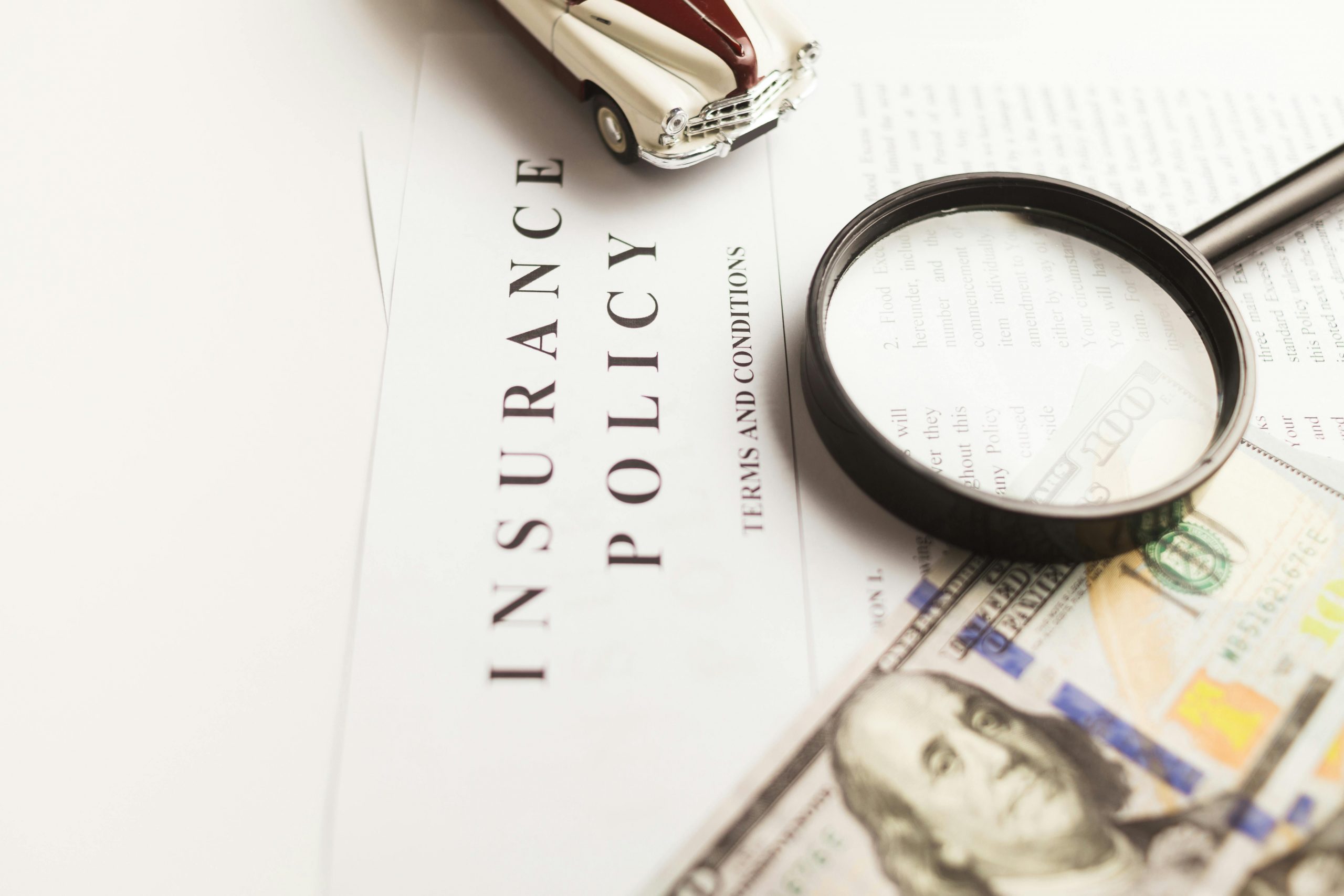 dollar bills, a magnifying glass, and a toy classic car placed on top of an insurance policy document